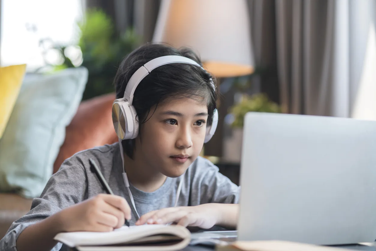 A boy wearing headphones, writing in a notebook while following an online lesson on a laptop
